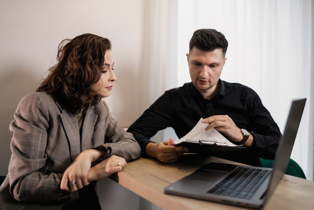 landlord-with-curly-brown-hair-in-a-brown-plaid-suit-reviews-a-rental-agreement-with-their-new-tenant-who-holds-a-clipboard-with-a-document-on-it