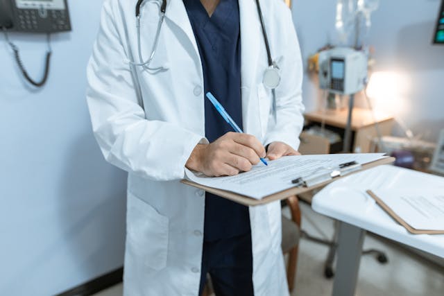 person-wearing-a-stethoscope-and-white-lab-coat-in-scrubs-writing-on-a-chart-clipped-on-a-clipboard